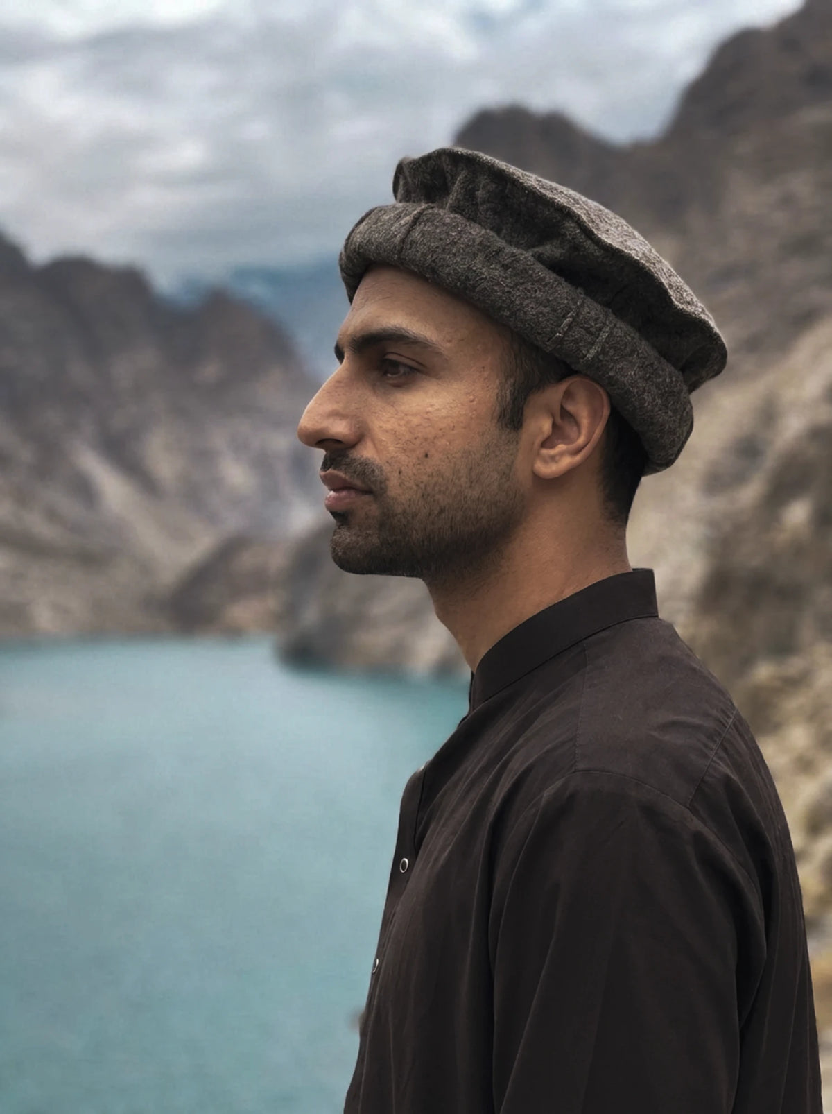 Hamza, founder of Montavita, in profile in a traditional pakol hat beside a mountain lake in Gilgit-Baltistan